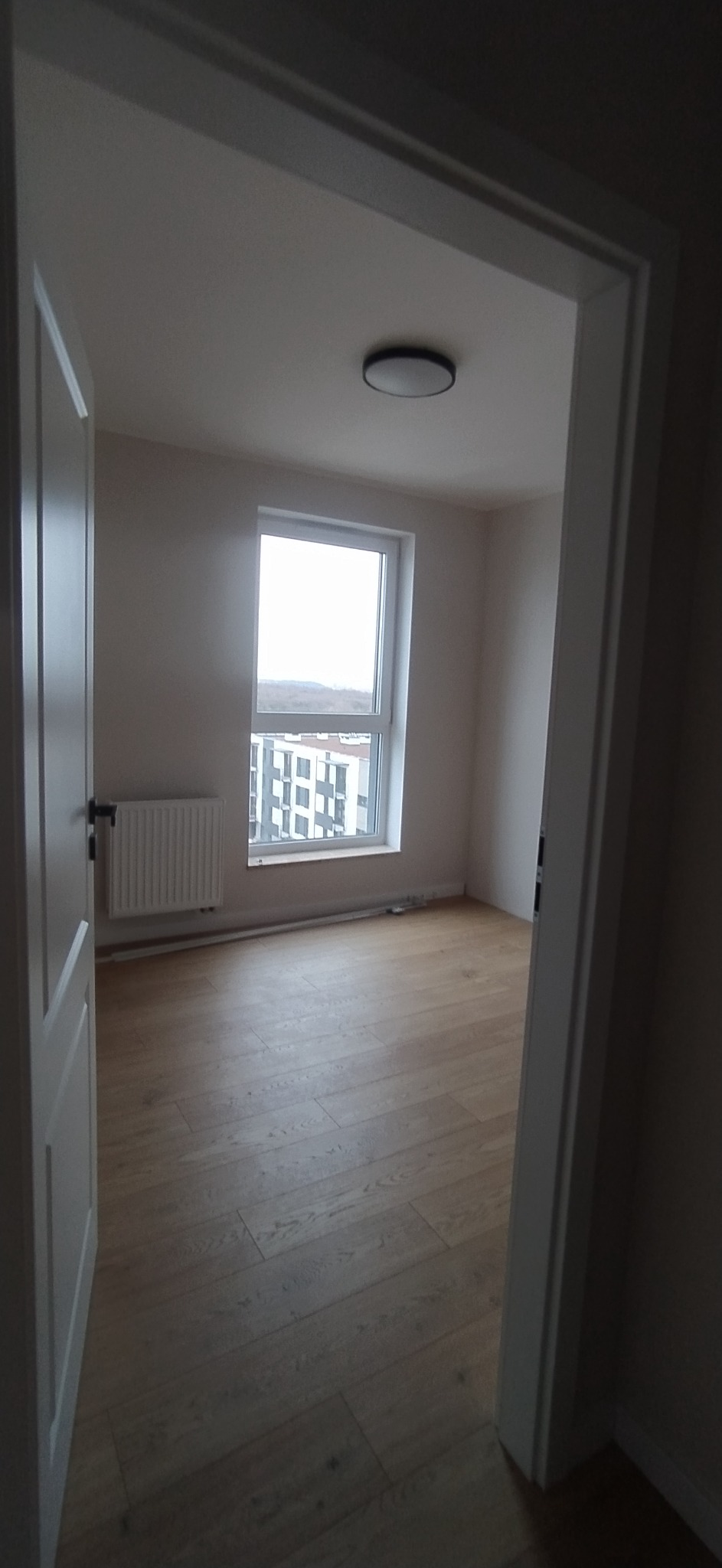 Jasne panele podłogowe ułożone w pokoju z widokiem na zabudowę miejską, widoczne przez okno; białe ściany i grzejnik pod oknem.