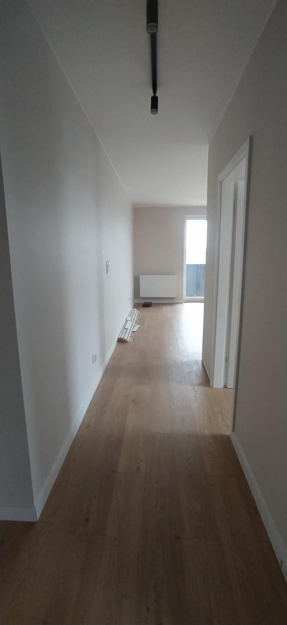 Korytarz po remoncie z jasnymi panelami podłogowymi, świeżo pomalowanymi ścianami w odcieniach szarości i białymi listwami przypodłogowymi. W tle widoczny grzejnik i okno balkonowe.