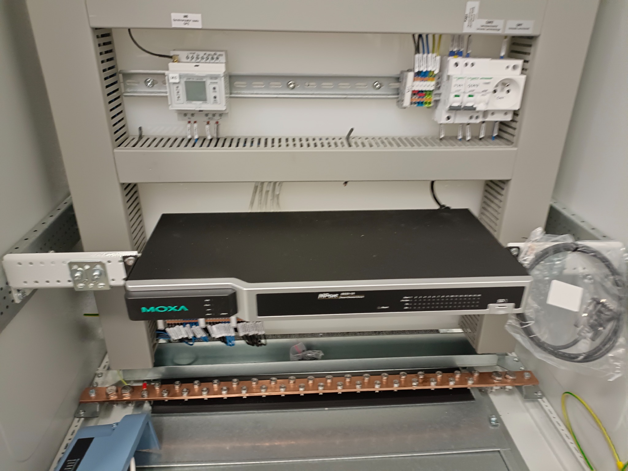 Wnętrze szafy elektrycznej z zainstalowanym urządzeniem MOXA, widoczne liczniki, bezpieczniki i listwa uziemiająca z przewodami.