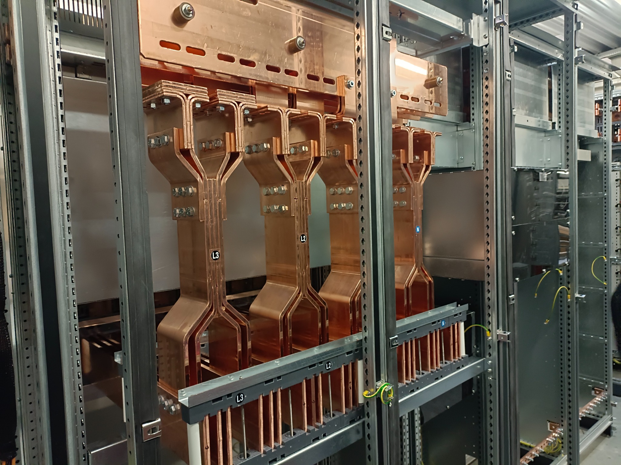 Miedziane szyny zbiorcze w rozdzielnicy elektrycznej, widoczne oznaczenia faz L2 i L3, elementy metalowej konstrukcji wsporczej.