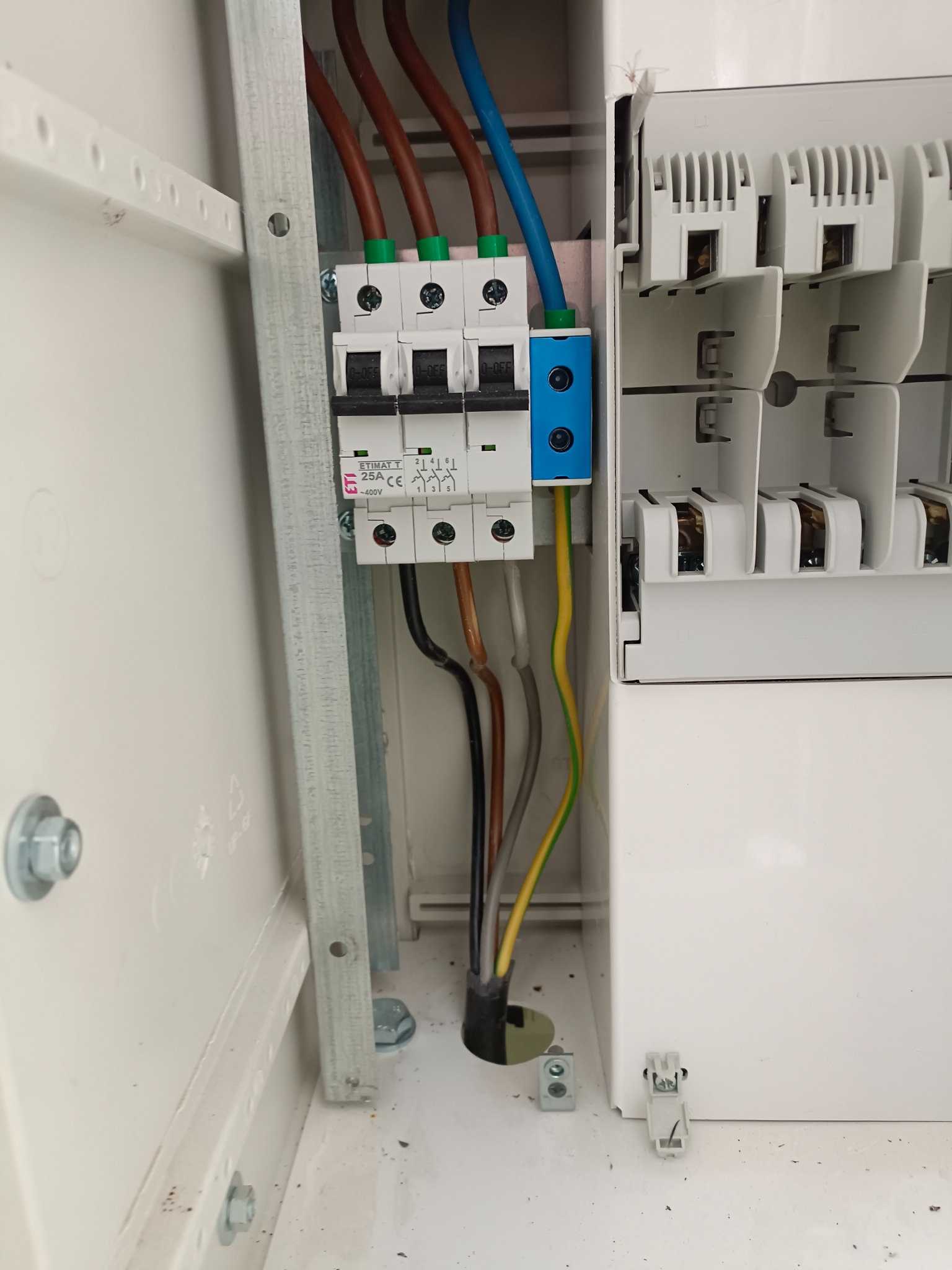 Otwarta skrzynka elektryczna z zainstalowanymi bezpiecznikami, widoczne przewody w różnych kolorach oraz metalowa szyna montażowa.