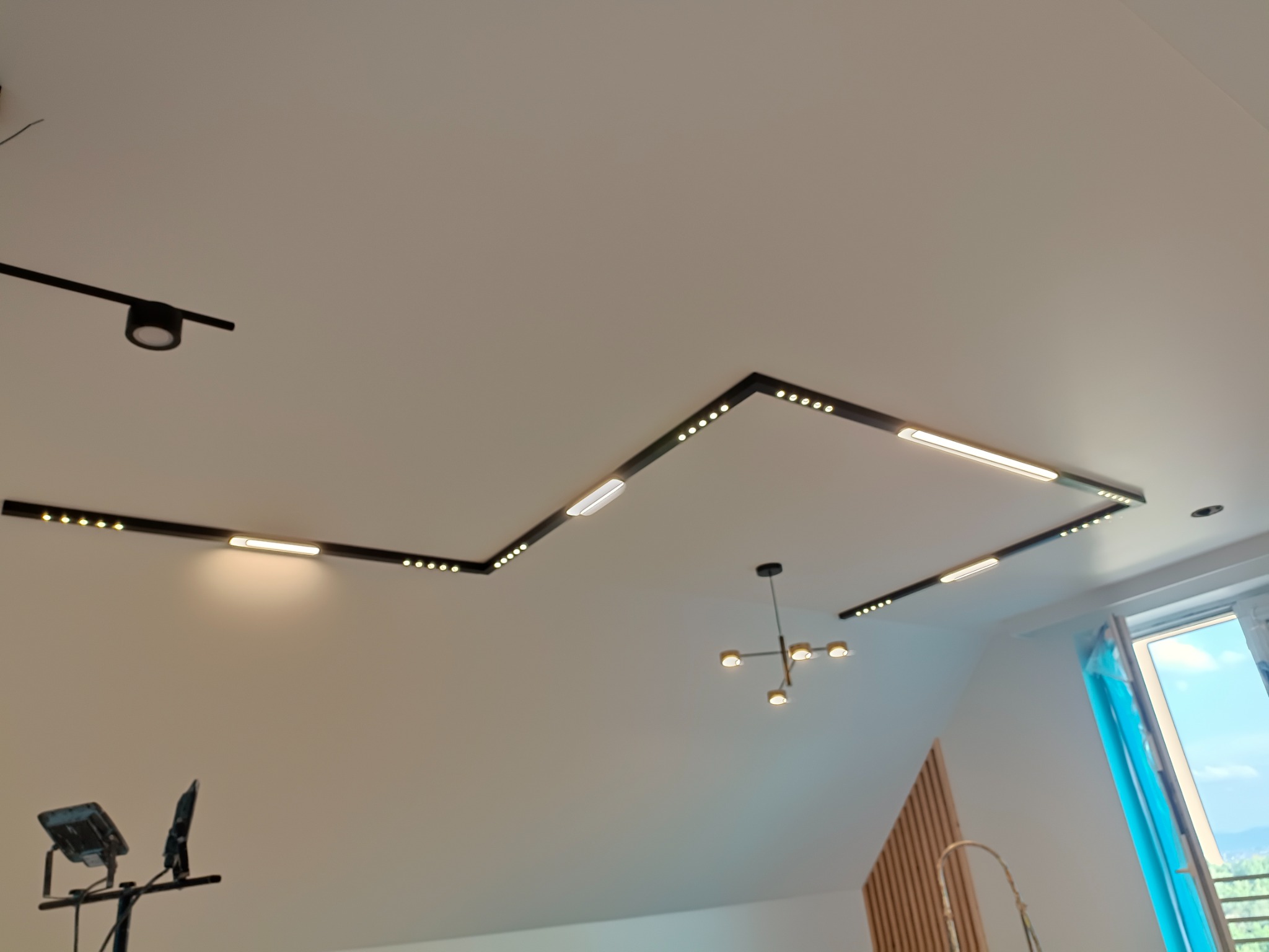 Czarna listwa oświetleniowa LED z punktowymi i liniowymi źródłami światła, zamontowana na białym suficie pod skosem, widoczna lampa wisząca i okno z turkusową zasłoną.