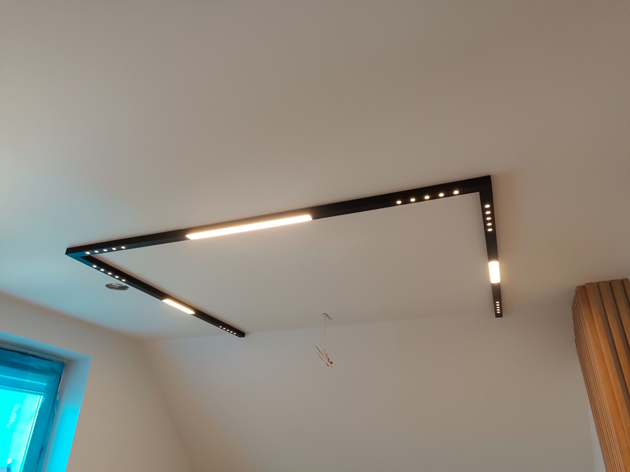 Czarna, geometryczna oprawa oświetleniowa LED na białym suficie, z widocznymi przewodami w centralnej części sufitu, częściowo widoczne okno i dekoracyjna ściana z drewnianych paneli w tle.