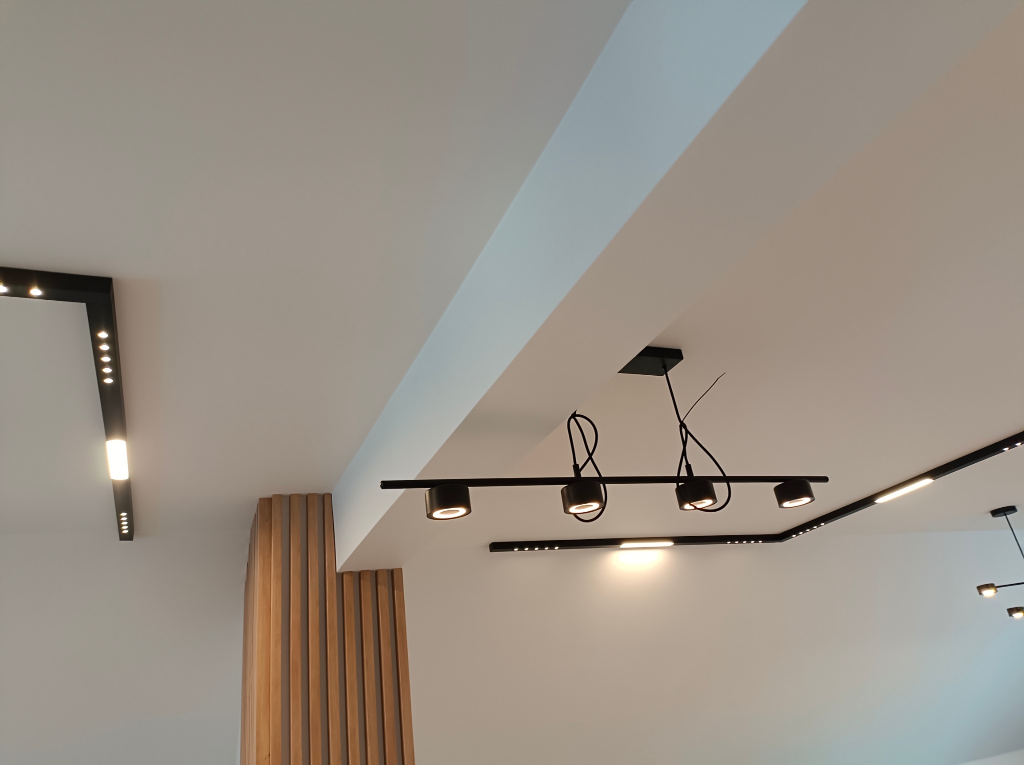 Nowoczesne oświetlenie sufitowe z czarnymi reflektorami i listwami LED, rozmieszczone w geometrycznym układzie na białym suficie, widoczne elementy dekoracyjne z drewnianych listew.