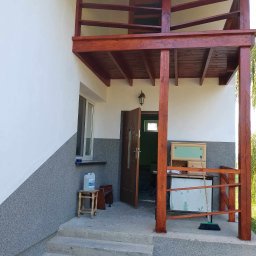 MATBUD - Dwukondygnacyjny, drewniany ganek z balustradą w kolorze brązu, przylegający do otynkowanego domu z szarą elewacją na dole i białą na górze, widoczne schody wejściowe, otwarte drzwi wejściowe...