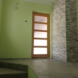 MATBUD - Wnętrze z zielonymi ścianami, drewnianymi drzwiami z przeszkleniami i fragmentem ściany wyłożonej dekoracyjną szarą cegłą, widoczne schody z ciemnej terakoty.