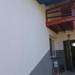 MATBUD - Ściana budynku z wykończoną elewacją: górna część biała, dolna szara, widoczny balkon z drewnianą balustradą, okno oraz drzwi wejściowe. Na zewnątrz stoi plastikowy kanister na małym stołku.