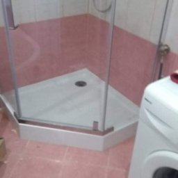 MATBUD - Narożna kabina prysznicowa z brodzikiem, widok z perspektywy podłogi, obok pralka, różowe płytki na podłodze i ścianach.