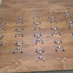 MATBUD - Układanie drewnopodobnych płytek podłogowych wewnątrz pomieszczenia, z widocznymi krzyżykami dystansowymi i klinami poziomującymi, przed fugowaniem.