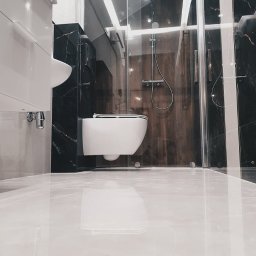 MATBUD - Nowoczesna łazienka z kabiną prysznicową ze szklanymi drzwiami, wiszącą miską WC i umywalką, wykończona płytkami imitującymi czarny marmur z białymi żyłami i błyszczącymi białymi płytkami podłogowymi.