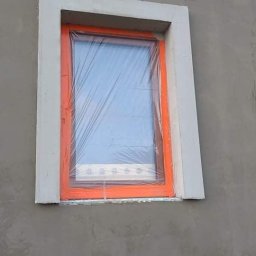 MATBUD - Okna zabezpieczone folią malarską podczas prac elewacyjnych; świeżo otynkowana ściana zewnętrzna z obramowaniem okiennym w kolorze szarym i pomarańczowym.