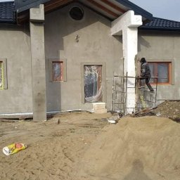 MATBUD - Pracownik na rusztowaniu wykańcza elewację domu jednorodzinnego w trakcie budowy, widoczny fragment ściany z oknami i drzwiami zabezpieczonymi folią, oraz sterta piasku na pierwszym planie.