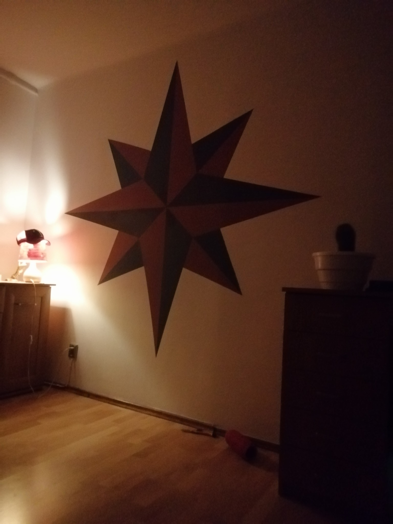 Duża, ręcznie malowana gwiazda w odcieniach czerwieni i szarości na ścianie w pokoju, oświetlona ciepłym światłem lampki nocnej. W tle widoczny drewniany parkiet i fragment mebla.