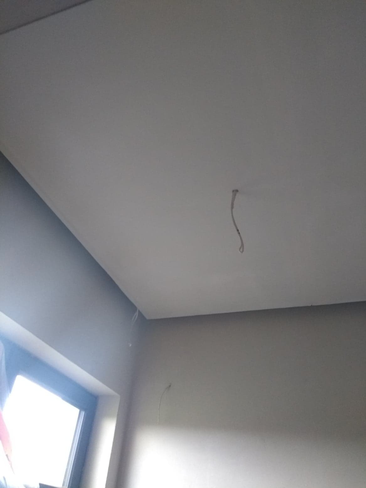 Wnętrze pokoju po malowaniu ścian i sufitu na odcienie szarości i bieli, widoczne kable wystające z sufitu, okno z jasnym światłem wpadającym do pomieszczenia.