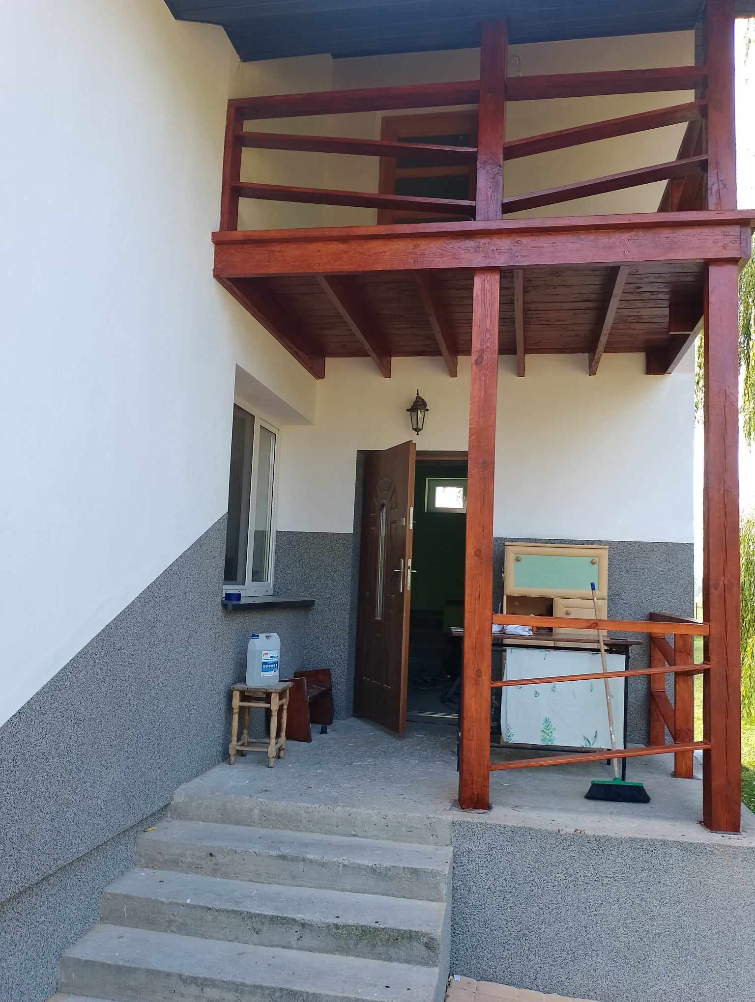 Dwukondygnacyjny, drewniany ganek z balustradą w kolorze brązu, przylegający do otynkowanego domu z szarą elewacją na dole i białą na górze, widoczne schody wejściowe, otwarte drzwi wejściowe...