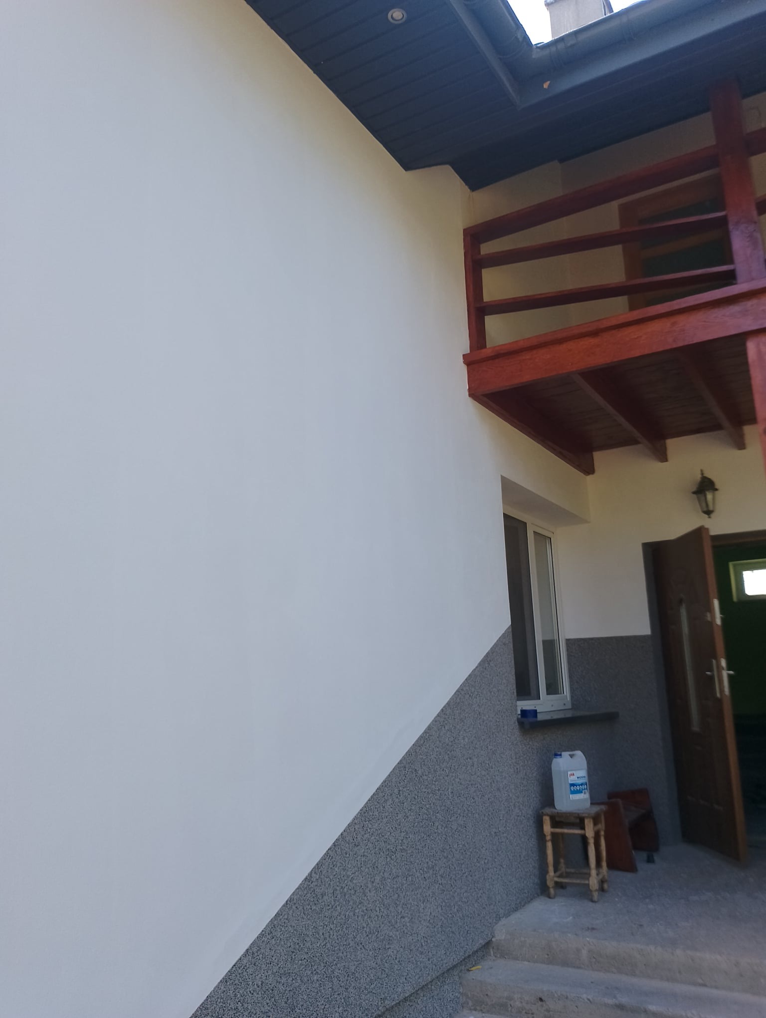 Ściana budynku z wykończoną elewacją: górna część biała, dolna szara, widoczny balkon z drewnianą balustradą, okno oraz drzwi wejściowe. Na zewnątrz stoi plastikowy kanister na małym stołku.