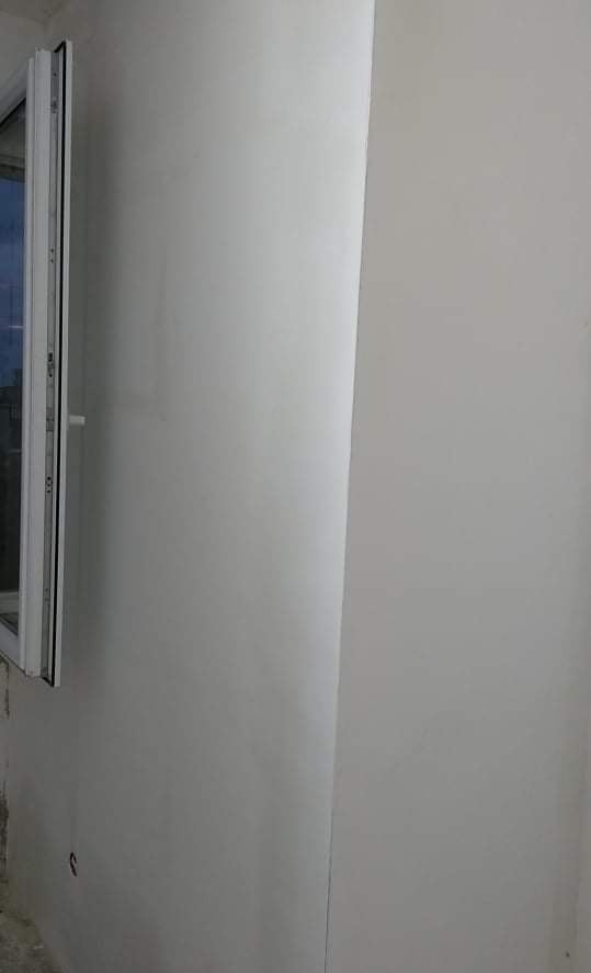 Świeżo pomalowana ściana w odcieniach bieli z widocznym fragmentem okna PCV po lewej stronie, ukazująca gładką powierzchnię po remoncie.