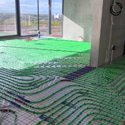 Bauinstalacje Witold Wałaszek - Zielone rury ogrzewania podłogowego ułożone na siatce w surowym wnętrzu z dużym oknem i betonowym słupem. Widok na krajobraz za oknem.