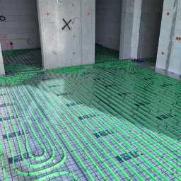 Bauinstalacje Witold Wałaszek - Zielone rury ogrzewania podłogowego ułożone na folii w surowym wnętrzu z betonowymi ścianami i otworami drzwiowymi. Widok z góry, równomierne oświetlenie.