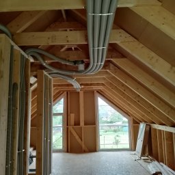 Bauinstalacje Witold Wałaszek - Montaż instalacji wentylacyjnej z szarymi rurami w surowym wnętrzu poddasza z drewnianą konstrukcją i oknami. Widoczne elementy mocujące i fragment krajobrazu za oknem.