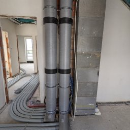 Bauinstalacje Witold Wałaszek - Szare, karbowane rury wentylacyjne z czarnymi opaskami, prowadzone wzdłuż ściany z bloczków betonowych, w surowym wnętrzu budynku w trakcie instalacji.