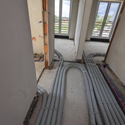 Bauinstalacje Witold Wałaszek - Szare rury wentylacyjne ułożone na podłodze w surowym wnętrzu, widoczne okna i fragmenty instalacji. Etap prac instalacyjnych w nowym budynku.