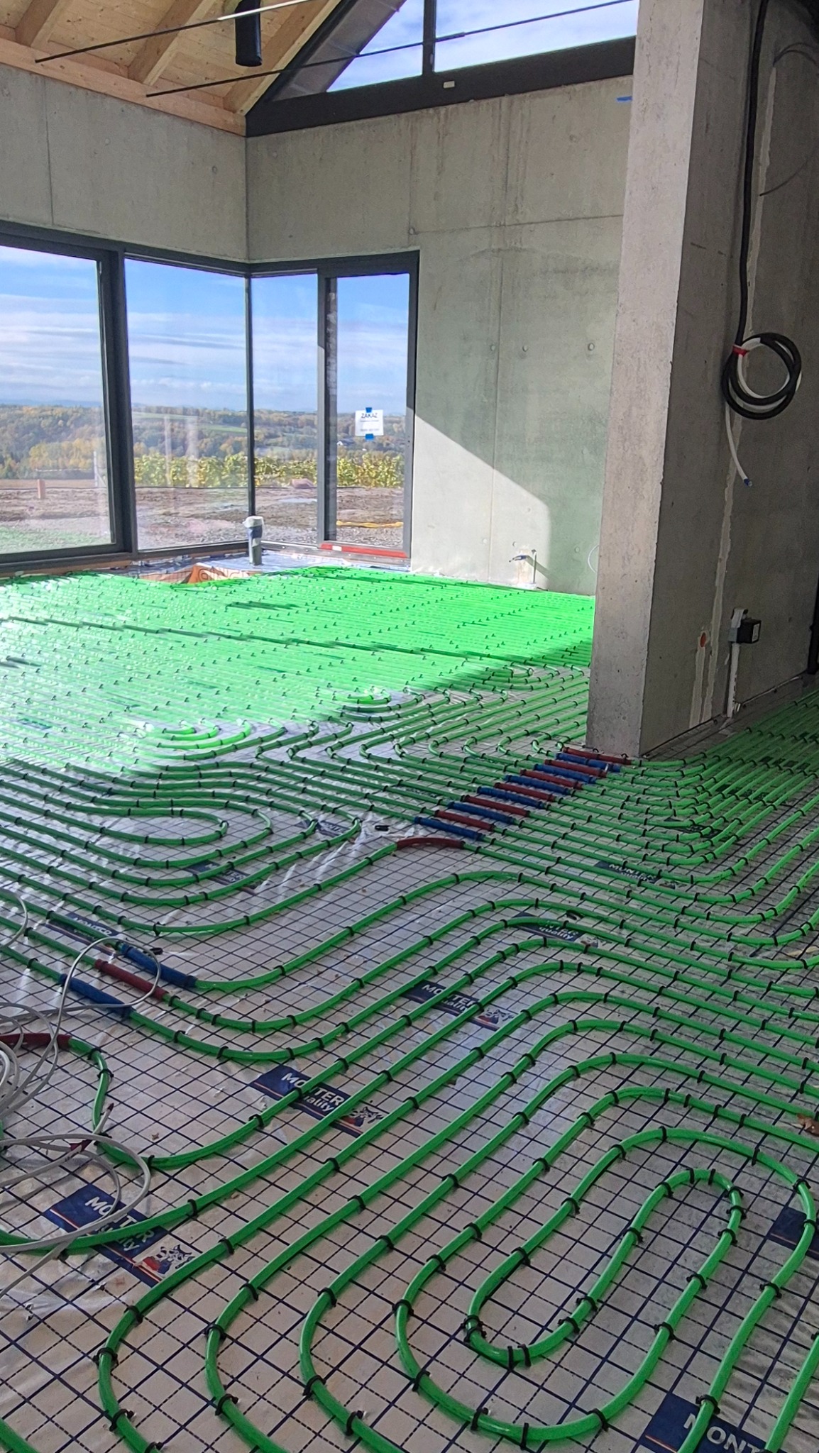 Zielone rury ogrzewania podłogowego ułożone na siatce w surowym wnętrzu z dużym oknem i betonowym słupem. Widok na krajobraz za oknem.