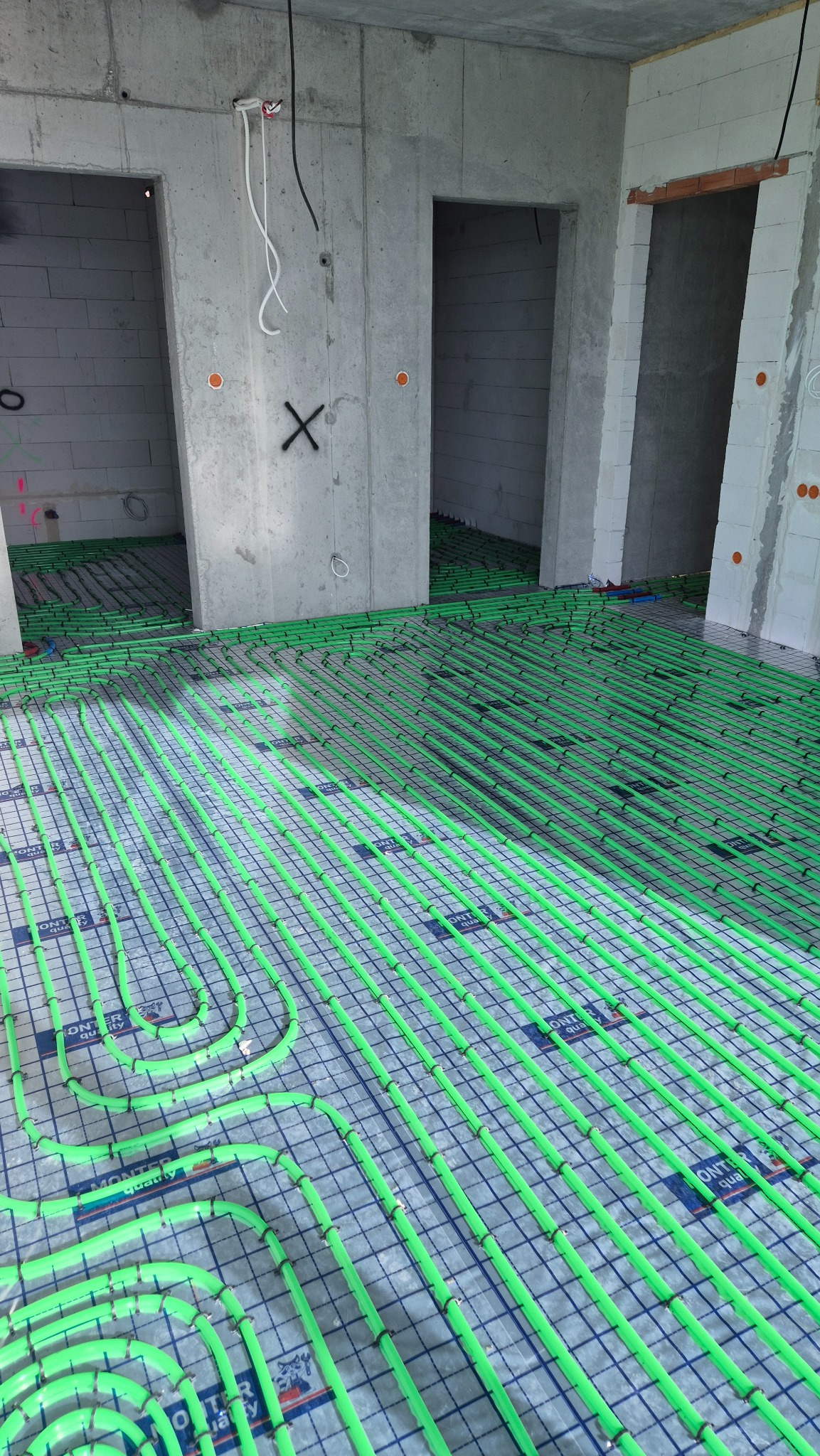 Zielone rury ogrzewania podłogowego ułożone na folii w surowym wnętrzu z betonowymi ścianami i otworami drzwiowymi. Widok z góry, równomierne oświetlenie.