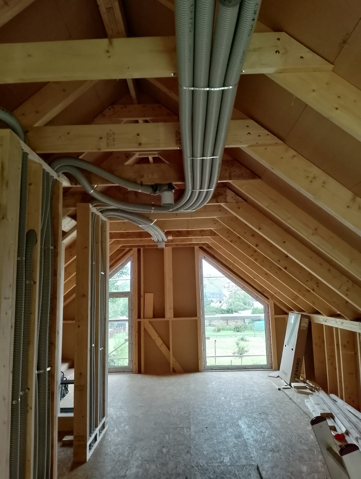 Montaż instalacji wentylacyjnej z szarymi rurami w surowym wnętrzu poddasza z drewnianą konstrukcją i oknami. Widoczne elementy mocujące i fragment krajobrazu za oknem.