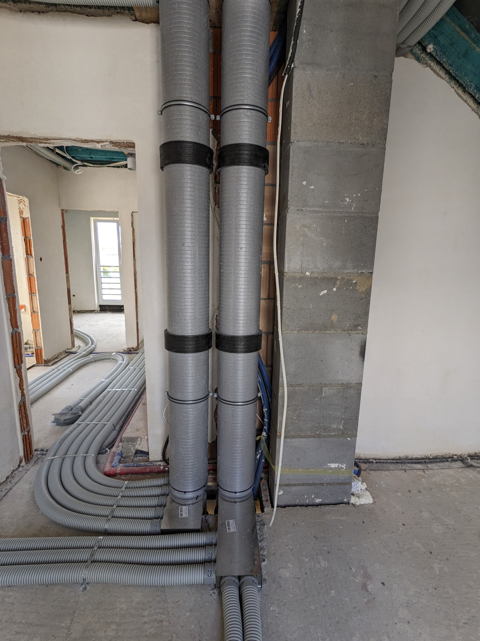 Szare, karbowane rury wentylacyjne z czarnymi opaskami, prowadzone wzdłuż ściany z bloczków betonowych, w surowym wnętrzu budynku w trakcie instalacji.