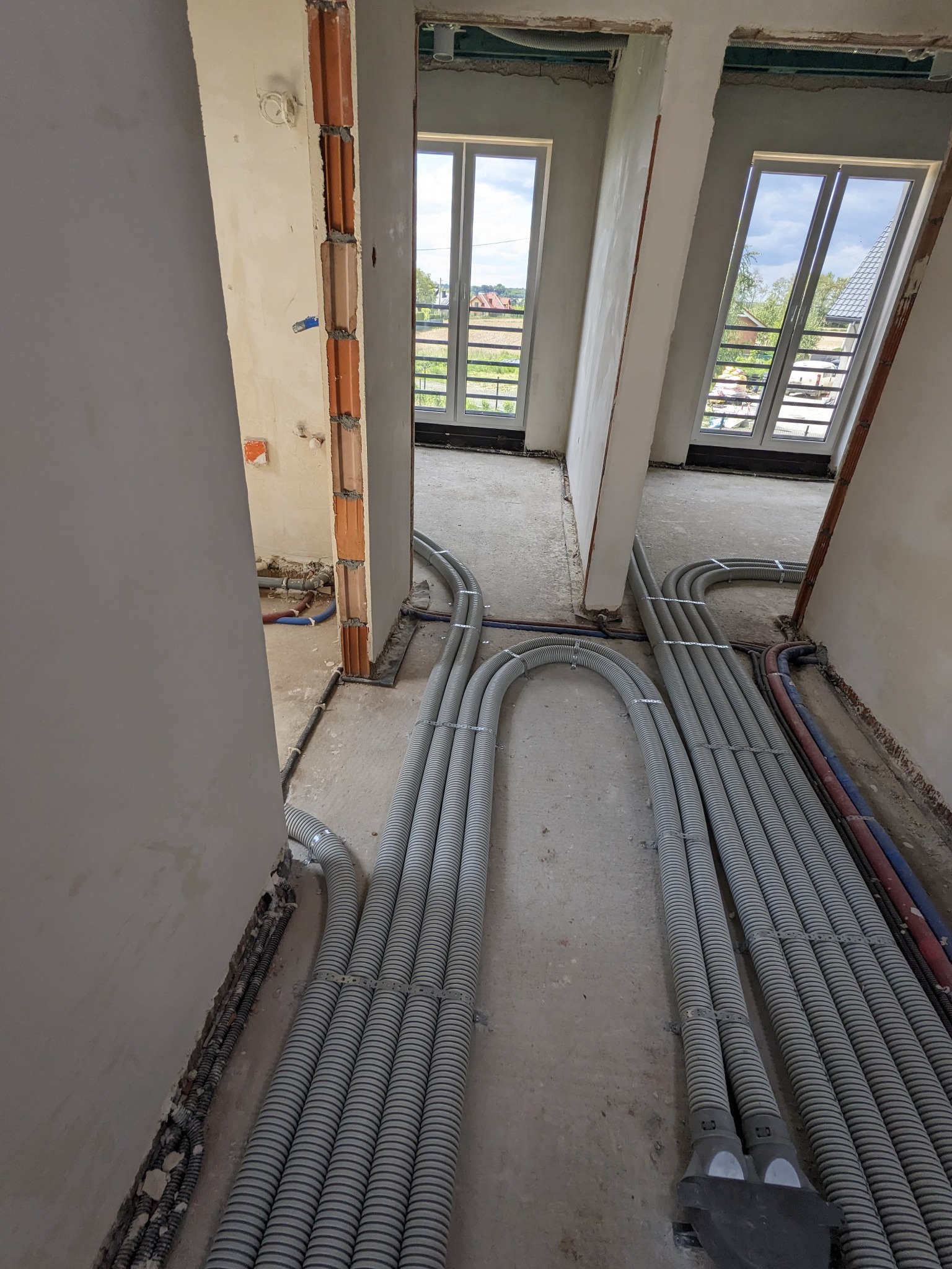 Szare rury wentylacyjne ułożone na podłodze w surowym wnętrzu, widoczne okna i fragmenty instalacji. Etap prac instalacyjnych w nowym budynku.