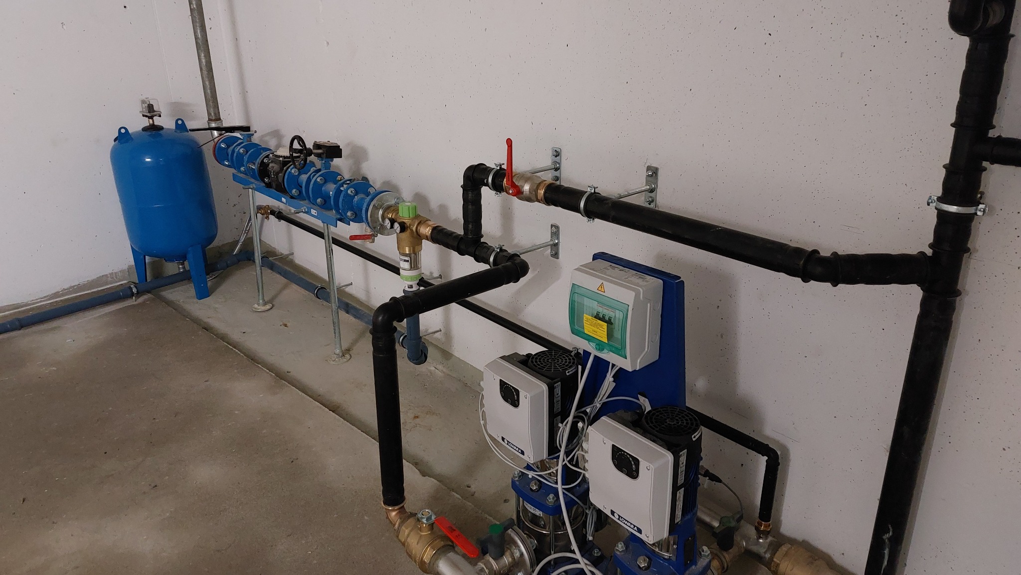 Złożona instalacja hydrauliczna z niebieskim zbiornikiem ciśnieniowym, czarnymi rurami i białymi urządzeniami sterującymi, zamontowana w pomieszczeniu technicznym.