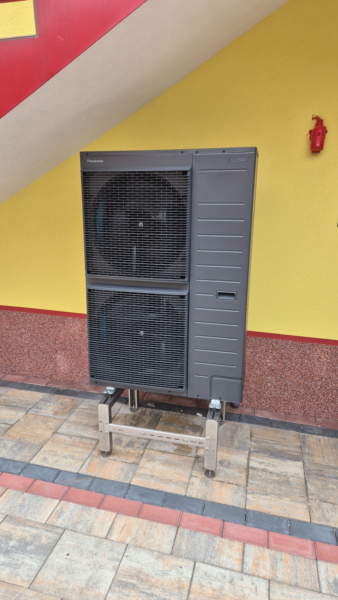 Szara pompa ciepła Panasonic Aquarea na metalowym stelażu, ustawiona na kostce brukowej przed żółtą ścianą budynku z czerwonymi akcentami. Widoczny zawór bezpieczeństwa.