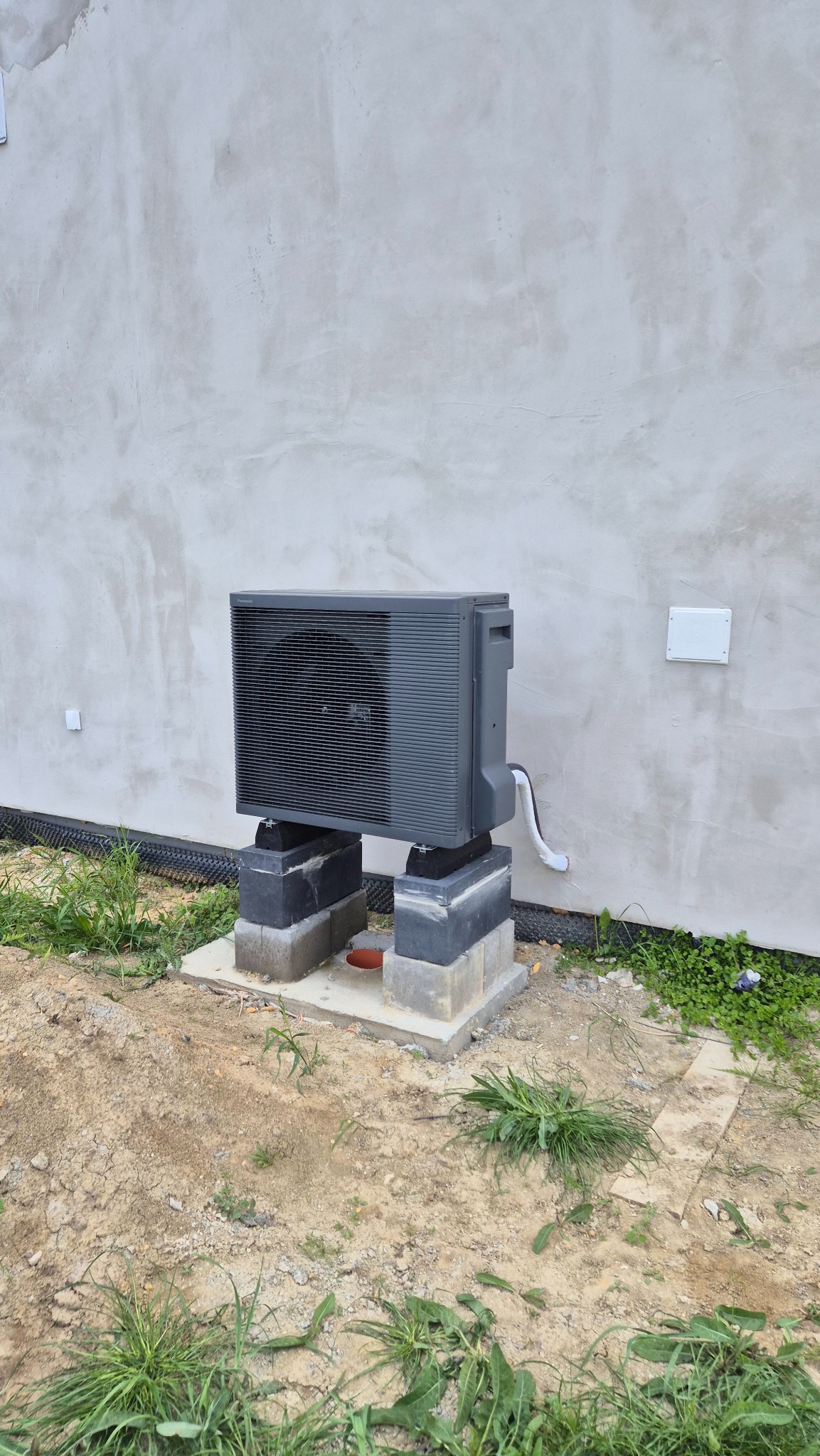 Szara pompa ciepła zamontowana na betonowych bloczkach przy ścianie budynku w trakcie wykończenia. Widoczne elementy instalacji i otoczenie z ziemią i trawą.