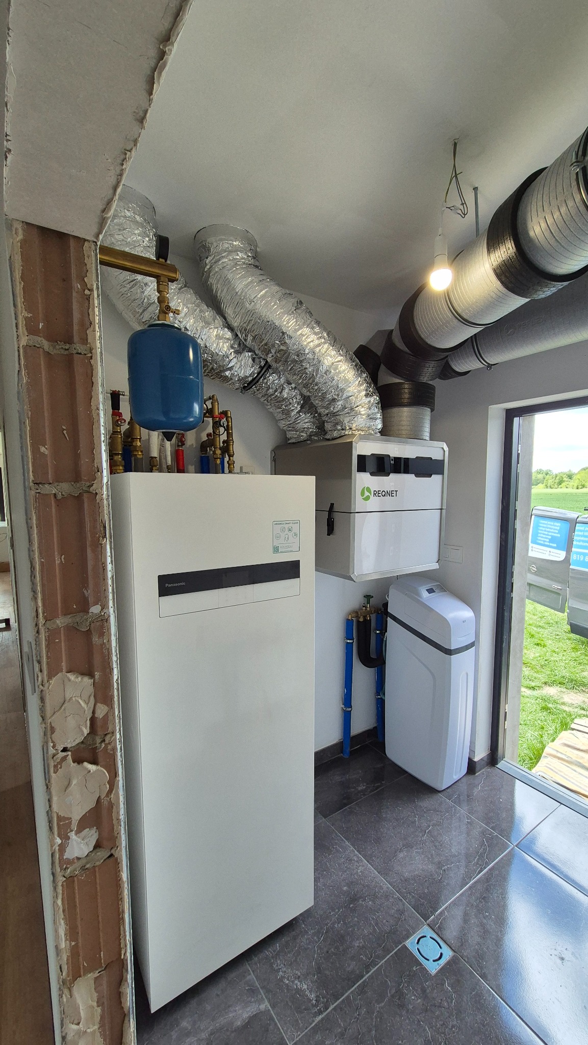 Nowoczesna kotłownia z rekuperacją: biały piec, srebrne rury wentylacyjne, niebieski zbiornik wyrównawczy i filtr wody. Szare płytki podłogowe.