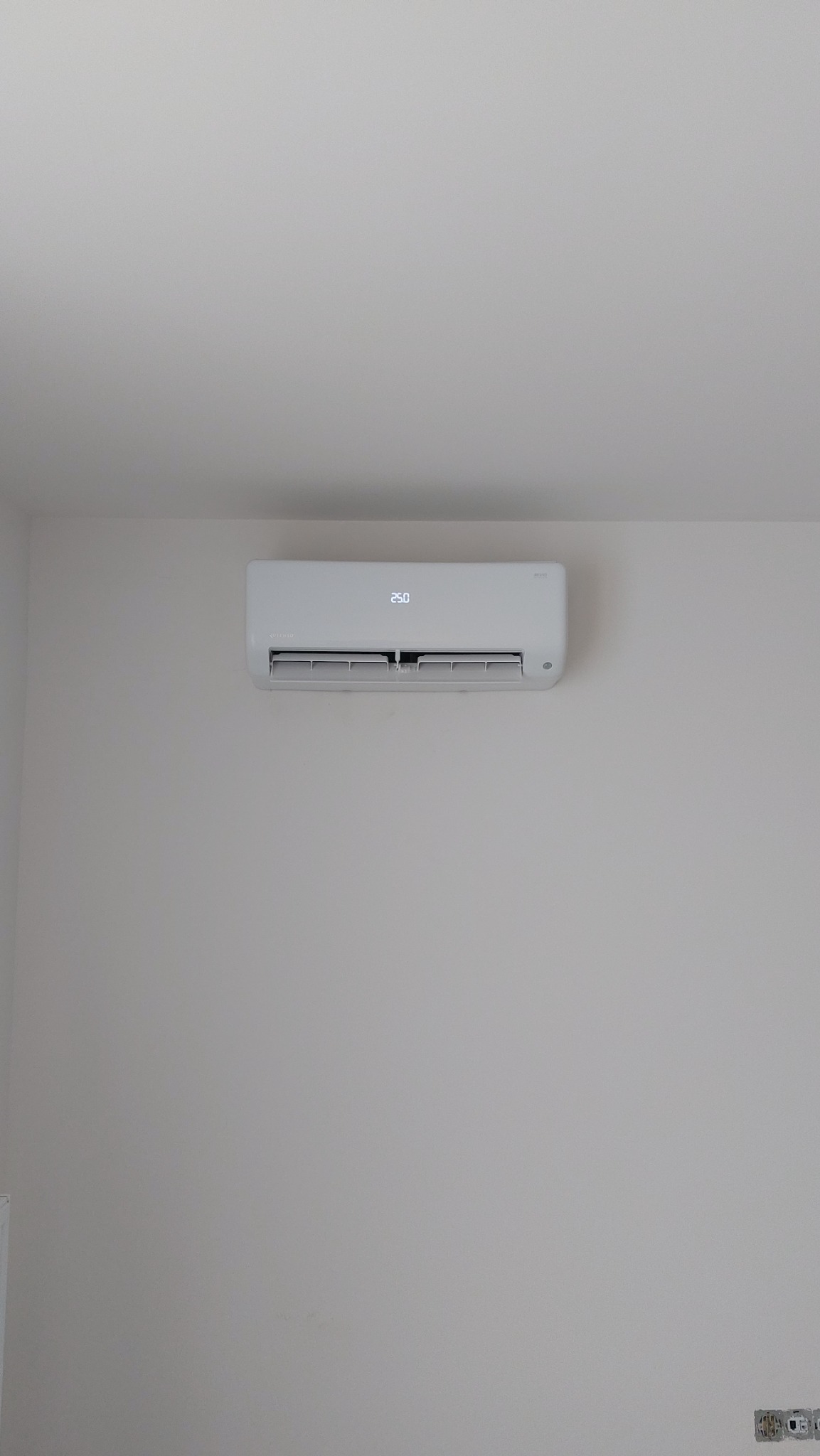 Biały klimatyzator ścienny z wyświetlaczem temperatury 25 stopni Celsjusza, zamontowany na gładkiej, jasnoszarej ścianie w minimalistycznym wnętrzu.