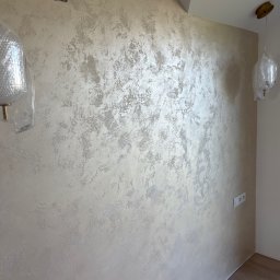 Smartdeco - Ściana z dekoracyjną, beżowo-srebrną strukturą, widoczne gniazdko elektryczne i fragment podłogi. Lampa wisząca w folii ochronnej. Otwór na kable w górnej części ściany.
