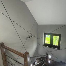 Smartdeco - Klatka schodowa po remoncie: szare ściany z czarnymi, geometrycznymi liniami, drewniane schody i balustrada ze stali nierdzewnej, okno z widokiem na zieleń.