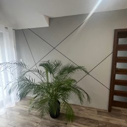Smartdeco - Jasne wnętrze z szarą ścianą zdobioną czarnymi liniami, drewnianą podłogą i drzwiami. W rogu stoi zielona roślina doniczkowa, a obok okna zwiewna, biała firanka.