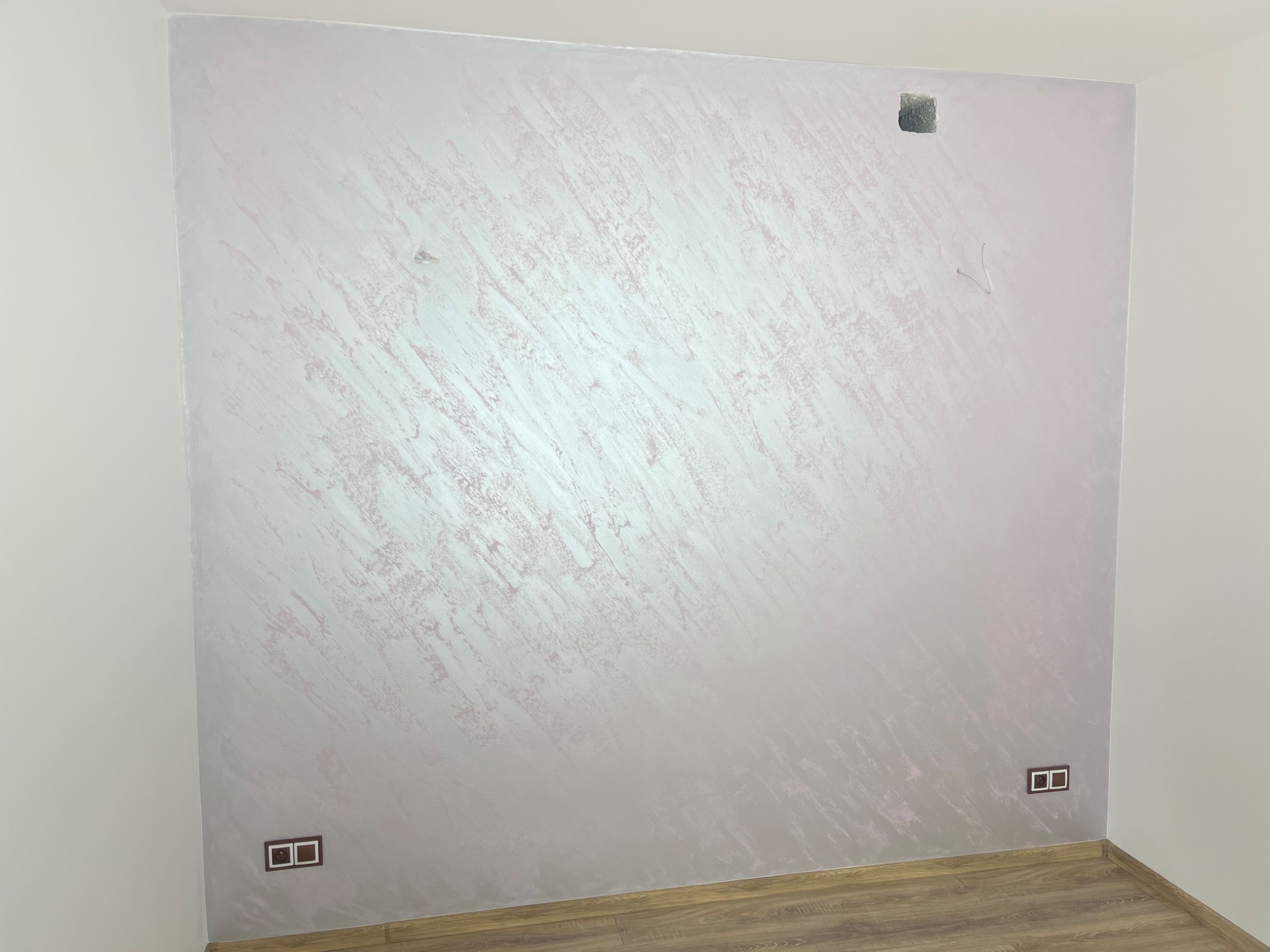 Ściana w pokoju po nałożeniu dekoracyjnej, perłowej farby z efektem smug, widoczne gniazdka elektryczne i listwa przypodłogowa.