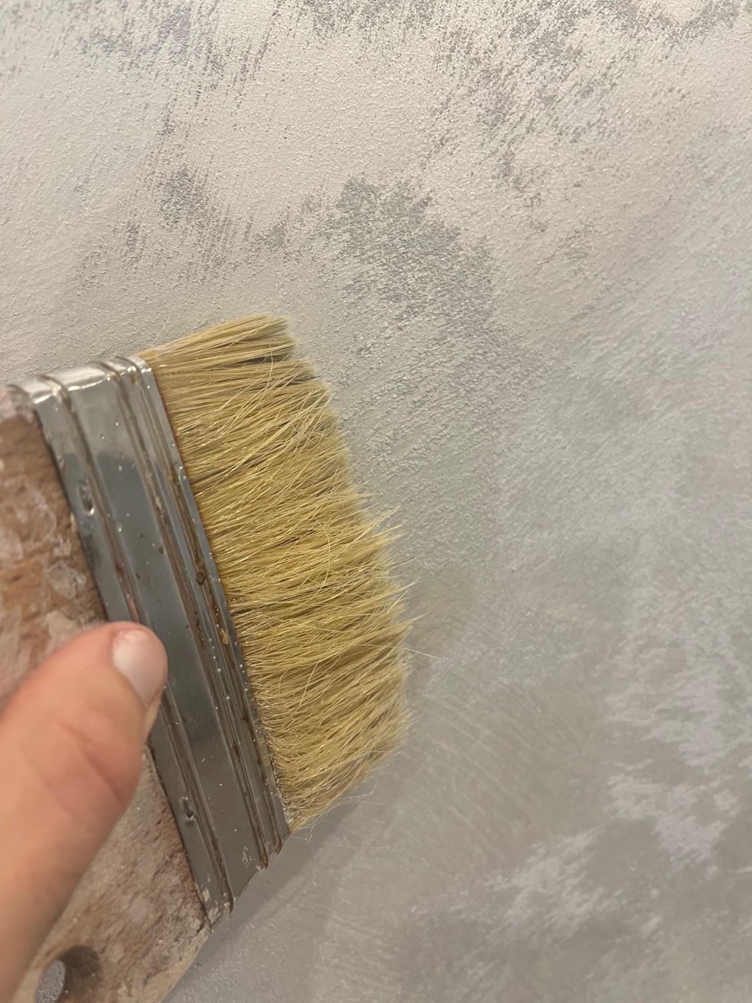 Nakładanie dekoracyjnej, szarej farby na ścianę za pomocą pędzla z naturalnym włosiem, widoczny fragment palca wskazującego.