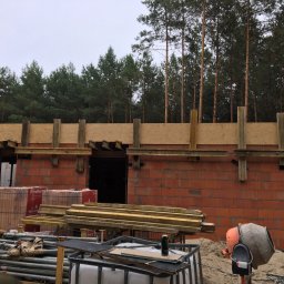 Gud-Bud - Wznoszenie muru z czerwonej cegły Porotherm na budowie domu, widoczne drewniane szalunki, palety z cegłami, betoniarka i las w tle.