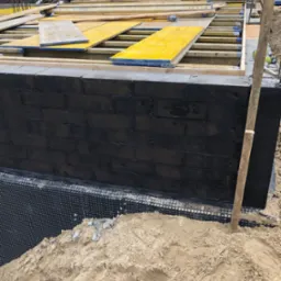 Częściowo ukończona ściana fundamentowa z czarną izolacją przeciwwodną, zabezpieczona folią kubełkową, z widocznym szalunkiem i deskami na g&oacute;rze, oraz stertą piasku u podstawy.