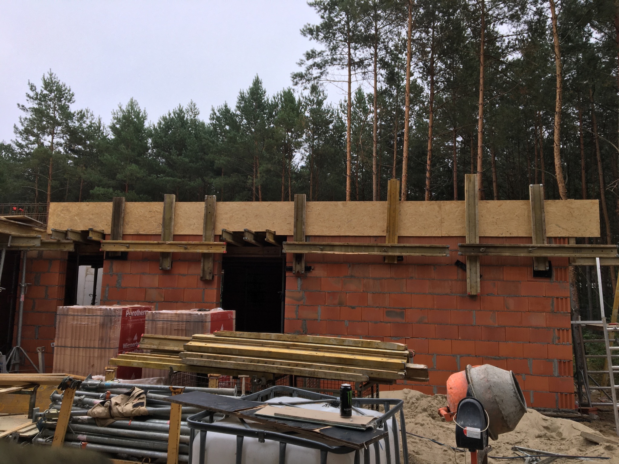 Wznoszenie muru z czerwonej cegły Porotherm na budowie domu, widoczne drewniane szalunki, palety z cegłami, betoniarka i las w tle.