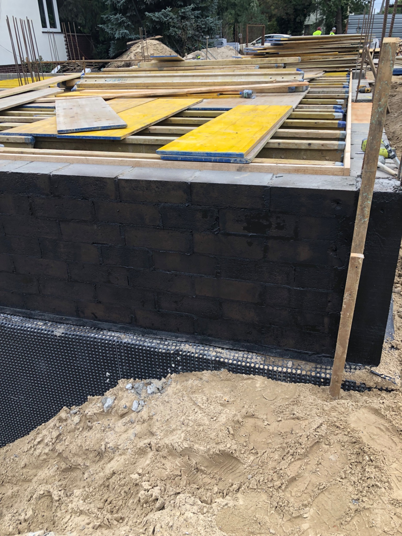 Częściowo ukończona ściana fundamentowa z czarną izolacją przeciwwodną, zabezpieczona folią kubełkową, z widocznym szalunkiem i deskami na górze, oraz stertą piasku u podstawy.