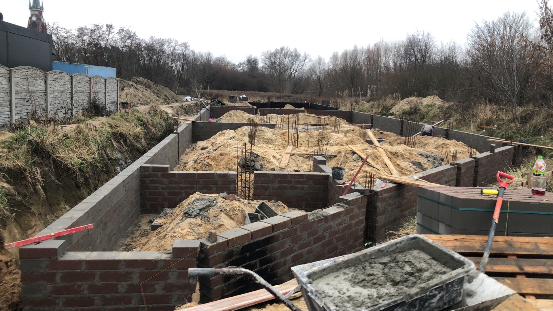 Wykop pod budynek z częściowo wymurowanymi ścianami fundamentowymi z czerwonej cegły, widoczne pręty zbrojeniowe i hałdy piasku wewnątrz, narzędzia murarskie na pierwszym planie, w tle drzewa...