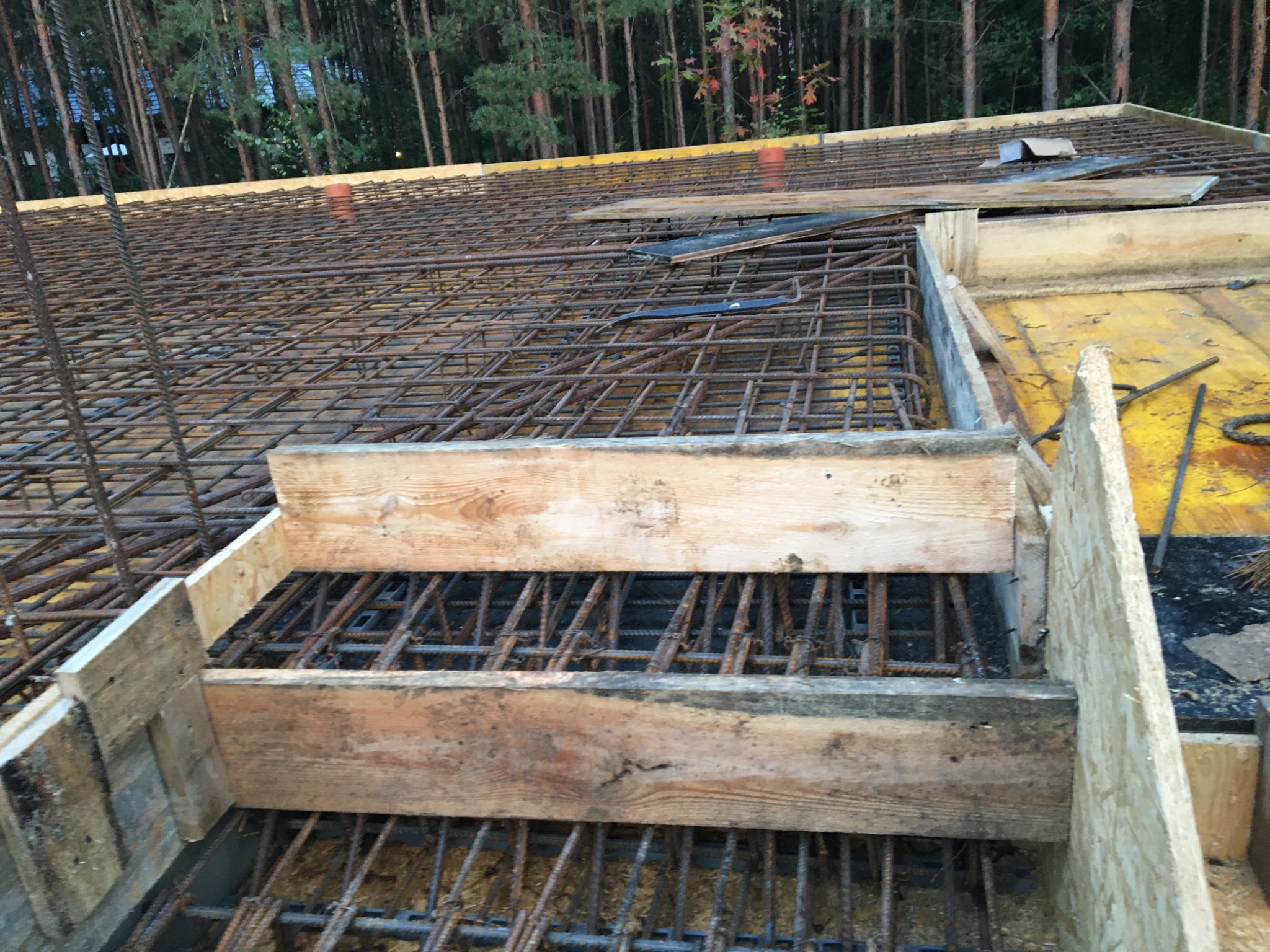 Zbrojenie płyty fundamentowej w deskowaniu z widocznymi prętami zbrojeniowymi i elementami drewnianymi, las w tle.