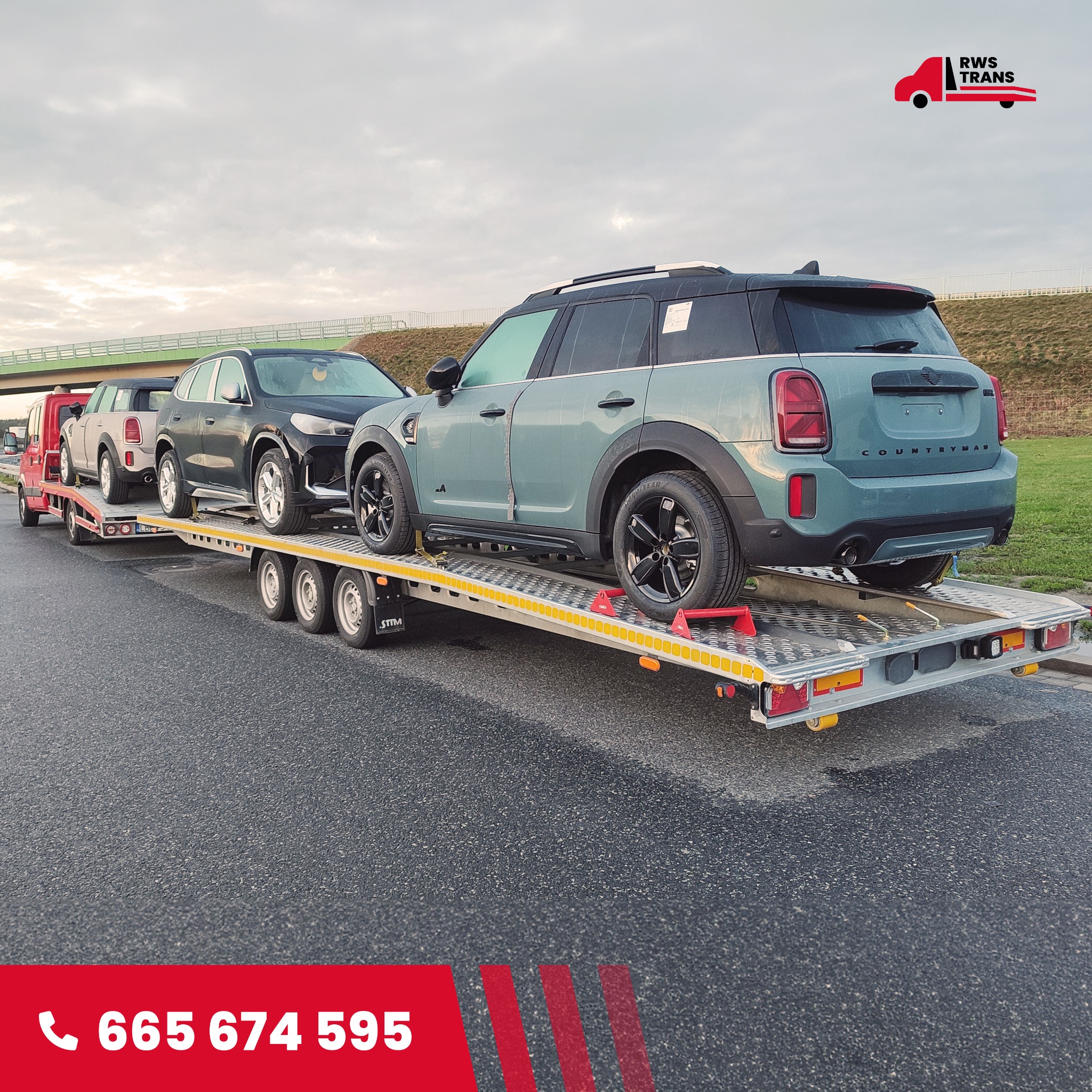 Niskozawieszona laweta z trzema samochodami osobowymi: zielony Mini Countryman, czarny BMW X1 i pickup, na tle pochmurnego nieba i wiaduktu.