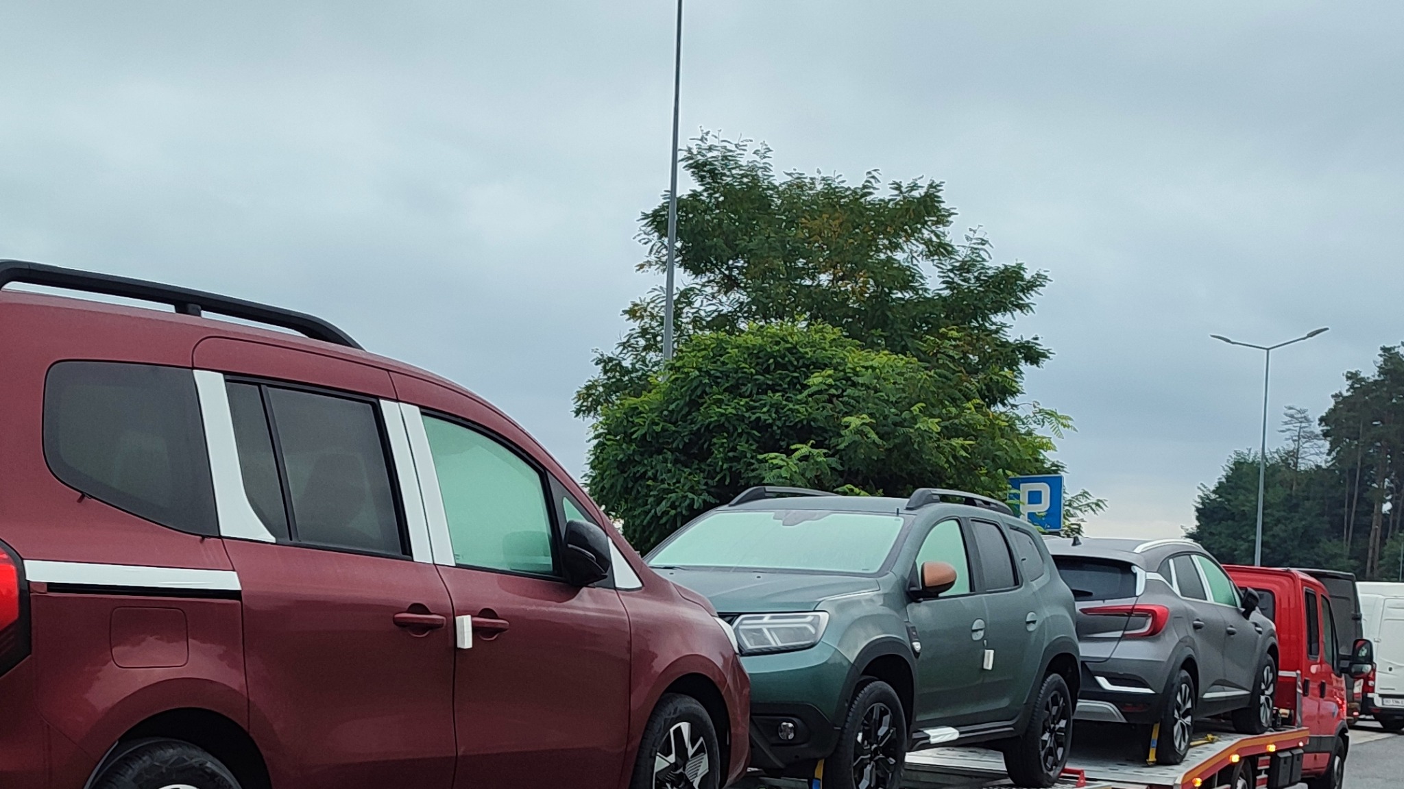 Czerwony, zielony i szary samochód osobowy na platformie, widok z boku, pochmurne niebo w tle.