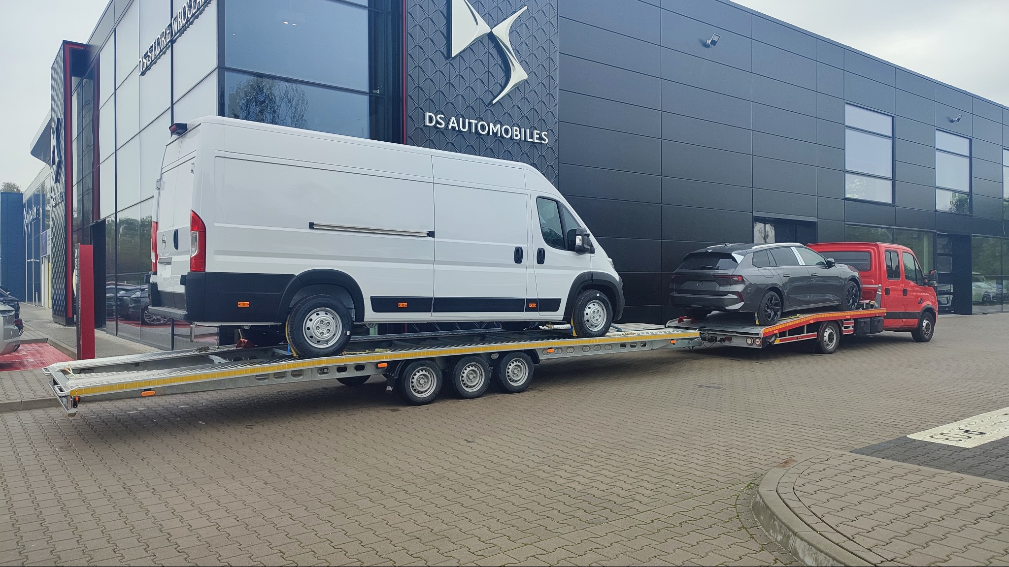 Dwa pojazdy na lawetach przed salonem DS Automobiles: biały furgon na dużej lawie i szary samochód osobowy na mniejszej, czerwonej lawecie.